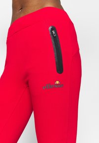 Rode sportieve broek met een slim fit, voorzien van een zwarte ritszak en een logo met een halve cirkelontwerp. Soepele textuur en rekbaar materiaal.