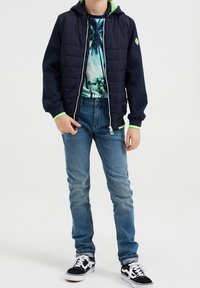 Modrá zateplená bunda s tmavě námořnick ým vnějším materiálem, zelenou podšívkou kapuce, zapínáním na zip, v kombinaci s tričkem s tropickým potiskem a modrými džíny.