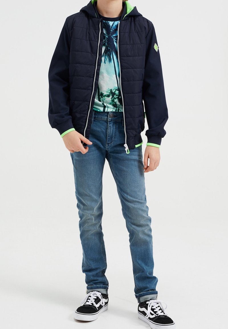 Modrá zateplená bunda s tmavě námořnickým vnějším materiálem, zelenou podšívkou kapuce, zapínáním na zip, v kombinaci s tričkem s tropickým potiskem a modrými džíny.