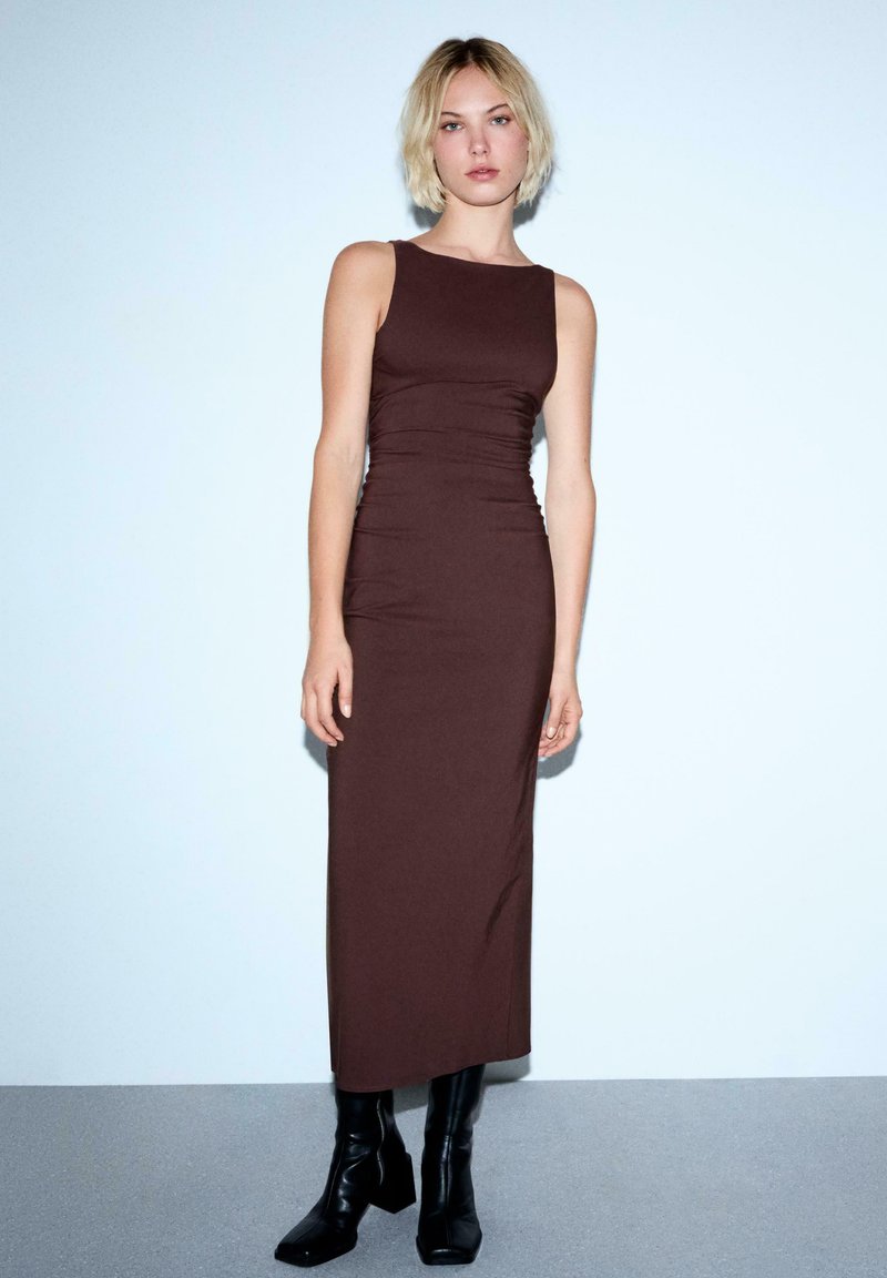 Rochie maxi, fără mâneci, de culoare maro, ajustată pe corp, cu un finisaj texturat și detalii pliate, asortată cu cizme negre până la glezne.