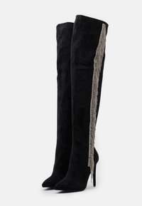Botas negras por encima de la rodilla en suave ante con punta afilada y tacón de aguja. Detalle fino con un fleco de acentos metálicos a lo largo del costado.