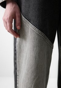 Prenda de denim con un diseño asimétrico que presenta secciones en gris oscuro y claro. Costuras visibles, detalles de bolsillo y textura suave.