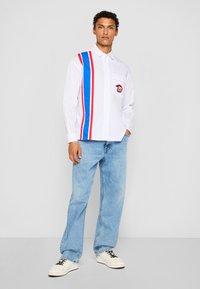 Hvid skjorte med knapper og røde og blå striber, med en brystlomme og logo-patch. Båret med lyseblå jeans og hvide sneakers.