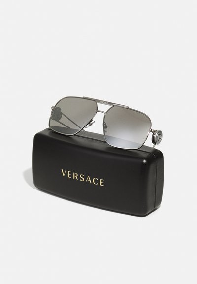 Ochelari de soare Versace cu un cadru argintiu, lentile gri cu degradé și accesorii decorative pe brațe, expuși deasupra unui toc negru.