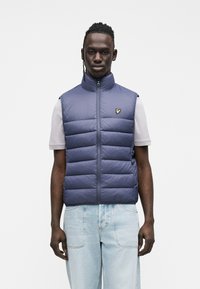 Vesta albastru închis căptușită, cu guler înalt, fermoar frontal și un logo brodat pe piept, purtată peste un tricou gri deschis și blugi din denim.