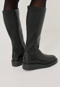 Fekete, térd fölé érő csizma sima bőrből és hálós panelekből, oldalsó cipzárral és lapos gumitalppal a stabilitásért.