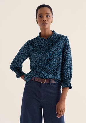 Femme portant un chemisier bleu à motifs avec col et poignets à volants, pantalon bleu foncé et ceinture marron sur fond uni.