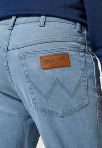 Los jeans de mezclilla azul claro presentan un parche de cuero marrón con el logo de Wrangler en el bolsillo trasero derecho y un detalle de costura en contraste.