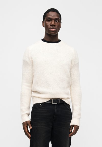 Pull à col rond en maille crème douce, doté de manches longues et de poignets côtelés, accompagné d'un jean en denim foncé et d'une ceinture noire.