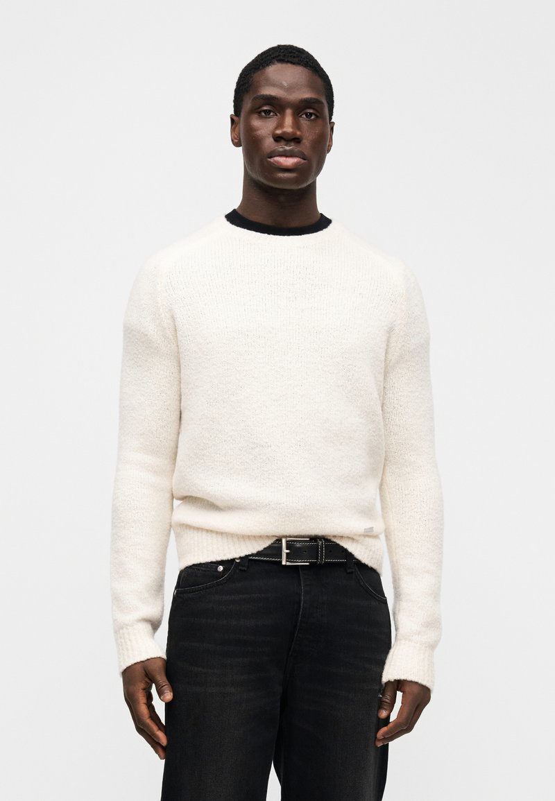 Crew neck sweater in soft cream knit, featuring long sleeves and ribbed cuffs, paired with dark denim jeans and a black belt.