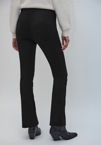 Pantalon noir évasé avec une texture lisse, doté de deux poches arrière et d'une taille haute, associé à des bottines noires à talons.