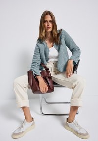 Cardigan di peluche azzurro chiaro sopra una t-shirt bianca, abbinato a pantaloni crema arrotolati e sneakers grigie. Una borsa bordeaux è tenuta in mano.