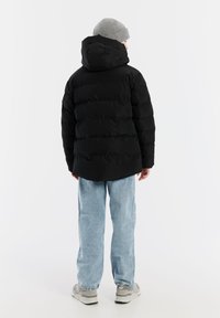 Zwarte gewatteerde jas met capuchon, voorzien van gestikte delen en een matte afwerking, gedragen over lichtblauwe jeans en grijze sneakers.