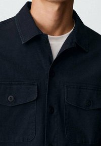 Ciemnoniebieska koszula z guzikami, wykonana z gładkiego materiału, z dwoma kieszeniami na piersi, kołnierzykiem i czarnymi guzikami. Prosty, zstructuredowany design.