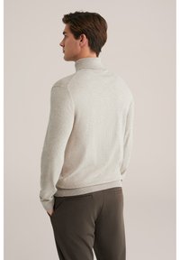 Pulover tricotat de culoare bej deschis, cu guler înalt, mansete și talie cu urmărire, purtat peste pantaloni închiși la culoare. Textură netedă și design ajustat.