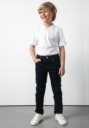 Biała koszulka polo z krótkim rękawem, granatowe dżinsy i białe sneakersy. Outfit ma klasyczny krój i casualowy styl.