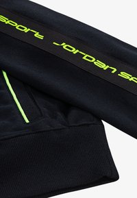 Răsărit dintr-un material negru de hanorac, cu un logo Jordan Sport de culoare verde neon scris cu caractere îndrăznețe și cusături de accent în aceeași nuanță neon.