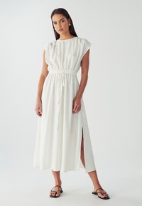 Bílá dlouhá šaty s krátkými rukávy, elastickým pasem a šňůrkou. Má boční rozparky a splývavý design. Nosené s černými sandály.