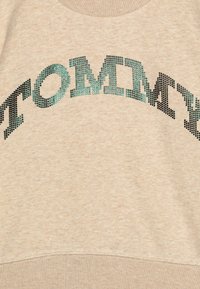 Beige mikina s texturovaným designem "TOMMY" v tyrkysových a černých kamínkách, s kulatým výstřihem a žebrovaným spodním lemem.