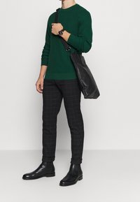 Homme portant un pull vert, un pantalon à carreaux noir, des bottes en cuir noir, un sac à bandoulière en cuir noir et une montre noire au poignet.