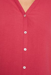 Maglietta rossa a V con una texture liscia, caratterizzata da bottoni bianchi lungo la parte anteriore. Il tessuto sembra leggero e informale.