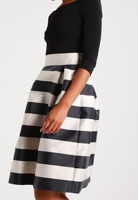 Haut à manches longues noir avec une jupe rayée contrastante en tissu noir et crème, présentant un design volumineux et plissé.