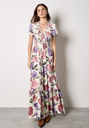 Donna che indossa un lungo abito floreale con rose rosa e viola, maniche a sbuffo, scollo a V, in piedi contro una parete chiara uniforme e un pavimento in cemento.