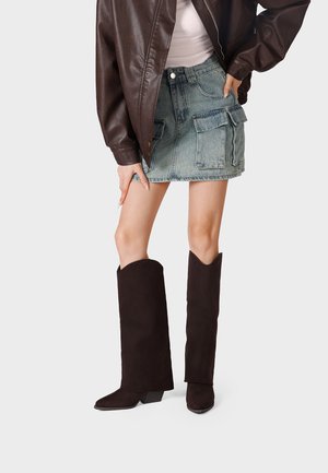 Persona che indossa una giacca di pelle marrone scuro, top bianco, minigonna di jeans con grandi tasche frontali e stivali marrone scuro alti fino al ginocchio, in piedi davanti a uno sfondo semplice.