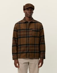 Veste en laine à carreaux bruns avec fermeture à bouton et poche. Portée avec un pantalon en velours côtelé beige et une casquette brune avec détail de logo.