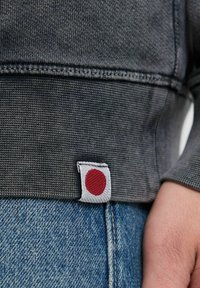 Grijze gebreide trui met een rood-wit geborduurd label aan de rand, gecombineerd met blauwe denim jeans. Close-up van de stof en de stiksels.