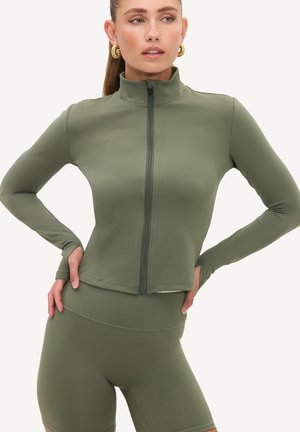Femme aux longs cheveux portant des boucles d'oreilles créoles dorées et une veste zippée vert olive assortie ainsi que des shorts de cycliste taille haute avec des trous pour les pouces.