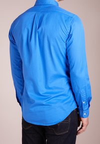 Mężczyzna w jasnoniebieskiej koszuli z długim rękawem i ciemnoniebieskich dżinsach, pokazany od tyłu na jednolitym jasnym tle.