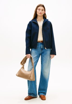 Νέα γυναίκα με σκούρο μπλε σακάκι, κρεμ πουλόβερ, ανοιχτό μπλε τζιν παντελόνι με φαρδιές γραμμές, καφέ μοκασίνια και κρατάει μια μπεζ τσάντα.