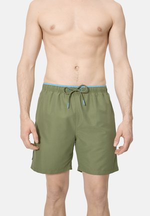 Hombre con pantalones cortos de baño verde oliva con cinturilla azul y cordón, de pie frente a un fondo liso.