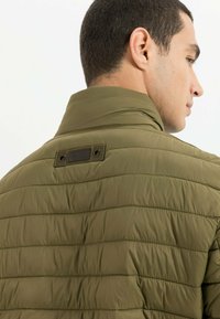 Olivengrøn puffjakke med vandret quiltning. Har en høj krave og et mærket tag på bagsiden, der viser en glat, blank tekstur.