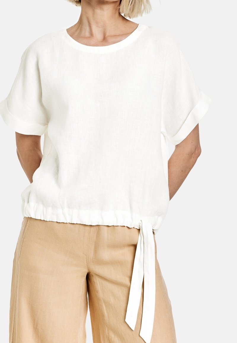 Haut en lin blanc à manches courtes, col rond et taille élastique froncée avec un détail noué. Associé à un pantalon beige clair.