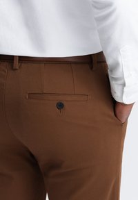 Braune maßgeschneiderte Hosen mit einer weichen Textur, ausgestattet mit einer Gesäßtasche und einem schwarzen Knopf. Tragen Sie dazu ein teilweise sichtbares weißes Hemd.