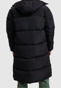 Svart puffjacka med huva, med ett quiltat mönster och förlängd silhuett. Slät textur och framträdande sömnadsdetaljer.