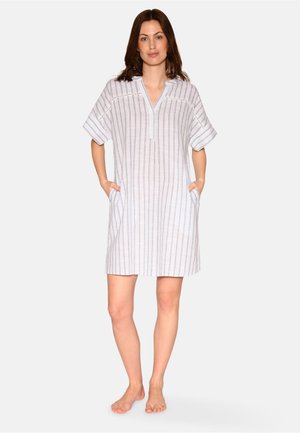 Femme debout pieds nus portant une robe à manches courtes, longueur genou, à rayures verticales avec des poches, sur un fond blanc uni.