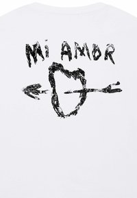 Witte T-shirt met een zwarte grafische afbeelding van een hart met een pijl en de tekst 'Mi Amor' erboven, wat een romantisch thema uitdrukt.