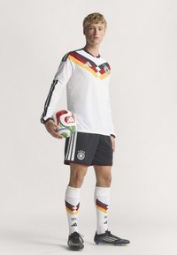 Witte lange mouwen jersey met een zigzagontwerp in rood, zwart en geel, gecombineerd met zwarte shorts, witte sokken met gekleurde accenten, en houdt een voetbal vast.