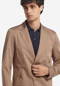 En beige blazer gjord av slät tyg, med snedkrage, enkel knappstängning och en bröstficka. Bärs över en marinblå polotröja.