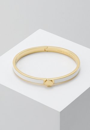 Gouden armband met witte emaille inzet en een gouden klavervormige sluiting, tentoongesteld op een wit geometrisch voetstuk.
