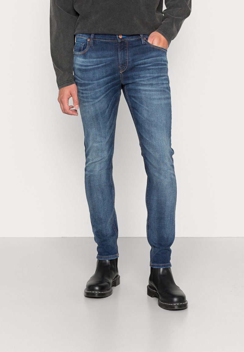 Jean skinny en denim bleu avec un design délavé, des coutures contrastantes et un style à cinq poches, associés à des bottines noires.