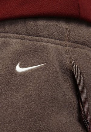 Bruine fleece joggers met een wit Nike-logo, elastische tailleband met drawstring en textuurdetail in de stof.