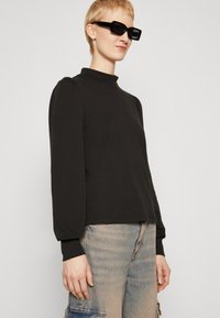 Black long-sleeve top featuring a high neckline and puffed sleeves, paired with light denim pants. The fabric appears smooth and lightweight.