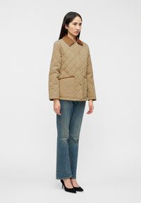 Μπεζ μπουφάν με σχέδιο καπιτονέ και καφέ γιακά και λεπτομέρειες στις τσέπες, με κουμπιά μπροστά, φορεμένο με μπλε τζιν καμπάνα και μαύρες γόβες.