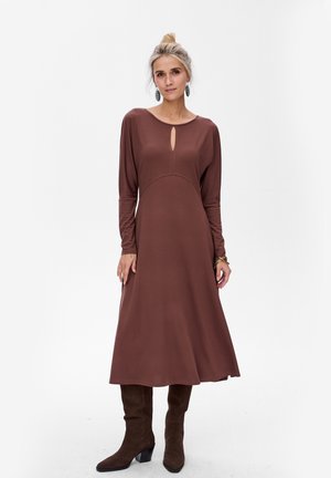 Robe longue marron à manches longues avec un décolleté en trou de serrure, corsage ajusté et jupe évasée. Portée avec des bottes marron jusqu'aux genoux et des accessoires simples.