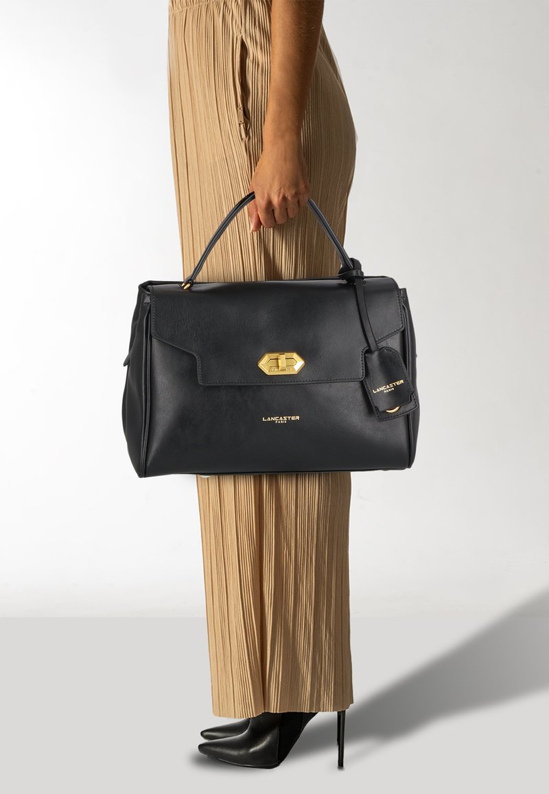Borsa a tracolla in pelle nera con chiusura in metallo dorato, design con pattina anteriore e etichetta attaccata. Abbinata a pantaloni beige plissettati e tacchi neri.