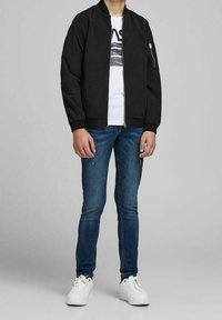 Czarna kurtka typu bomber na zamek, zestawiona z białym t-shirtem z grafiką i niebieskimi jeansami. Obuwie to białe trampki. Stylizacja casualowa.